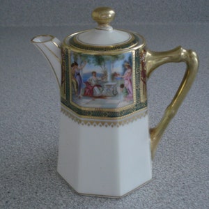 May include: A white porcelain creamer with a gold handle and lid. The creamer has a decorative scene of people in a garden painted on the side. The creamer is octagonal in shape.