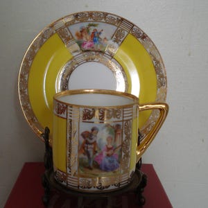 May include: A yellow and gold porcelain teacup and saucer set with a floral design and a couple in a romantic scene. The set is on a black metal stand.