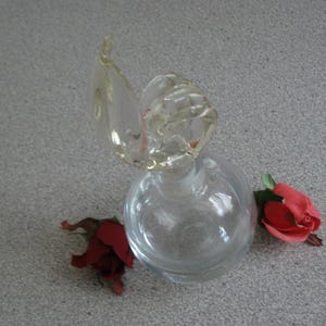 May include: A clear glass perfume bottle with a decorative stopper and two red roses. The bottle has a round base and a unique stopper design. The roses are a vibrant red color, adding a touch of elegance.