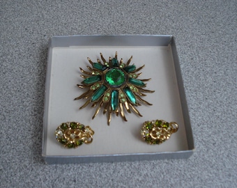 May include: A set of gold tone jewelry with green and clear rhinestones. The set includes a large sunburst brooch and a pair of earrings.