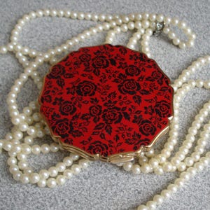 May include: A round compact mirror featuring a vibrant red background with a detailed black floral rose pattern and a scalloped gold-tone metal edge, resting atop a long strand of classic white faux pearls with a smooth, lustrous finish.