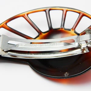 May include: A brown tortoiseshell hair clip with a fan-shaped design and a metal clasp. The clip has a silver metal bar with the words "Made in France" engraved on it.