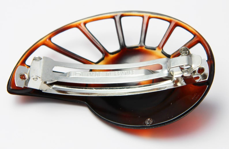 May include: A brown tortoiseshell hair clip with a fan-shaped design and a metal clasp. The clip has a silver metal bar with the words "Made in France" engraved on it.