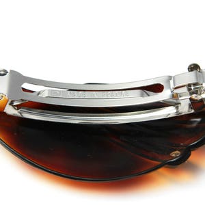 May include: A brown tortoiseshell hair clip with a silver metal clasp. The clip is shaped like a half-circle and has a textured surface.