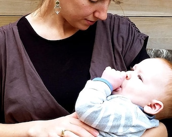 May include: A woman wearing a brown wrap top holds a baby in her arms. The baby is wearing a striped shirt and has a hand in their mouth. The woman is looking down at the baby. The background is a wooden wall with a sign that says "Je'ena design studio".