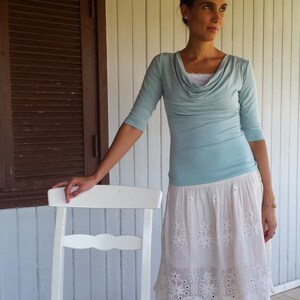 Puede incluir: Una mujer con una blusa azul claro de cuello de pico y una falda blanca de encaje con bordados florales. Est&aacute; de pie junto a una silla de madera blanca.