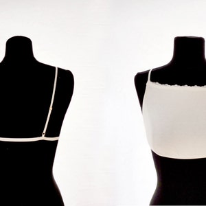Puede incluir: Bralette blanco con ribete de encaje en un maniquí negro. El bralette tiene tirantes finos y un diseño sencillo.