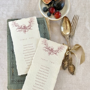 May include: Two table cards with a floral design and the names of guests for a wedding reception. The cards are on a linen tablecloth with a bowl of fruit and silverware.