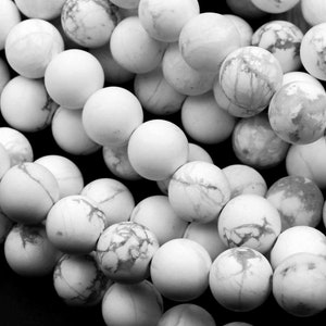 May include: A close-up of a pile of white howlite beads. The beads are round and have a smooth, polished finish. They are a light gray color with white veins running through them.
