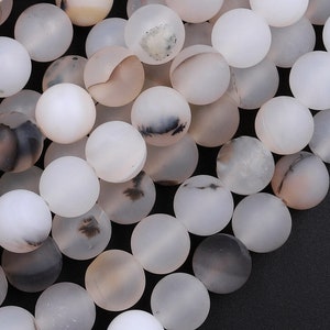 Puede incluir: Primer plano de una colección de cuentas redondas mate en tonos blanco, crema y marrón claro. Algunas cuentas tienen marcas grises y negras más oscuras. Las cuentas están ensartadas, lo que sugiere que son para hacer joyas.