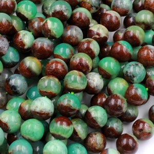 May include: A pile of spherical beads in varying shades of green, brown, and grey. The beads have a polished, glossy surface and look to be made of stone or a similar material. The beads are scattered, suggesting they are loose and ready for use in jewellery making or crafts.