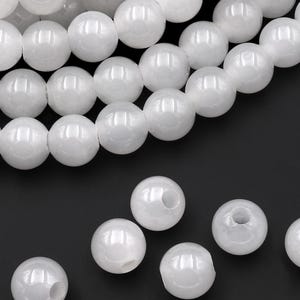 Peut inclure: Gros plan sur des perles blanches et rondes, enfilées et en vrac. Les perles ont une finition brillante et sont disposées sur un fond noir. Les perles sont adaptées à la fabrication de bijoux. Les perles mesurent environ 8 mm de diamètre.