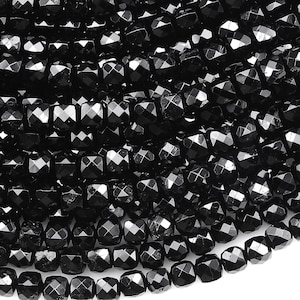 May include: Close-up of a collection of black, faceted beads. The beads are square-shaped with multiple facets, creating a sparkling effect. The beads are strung together, forming a dense, textured surface. The overall impression is of a dark, shimmering material.