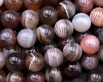 May include: A collection of round, polished beads in various shades of brown, gray, and cream. Some beads have striped patterns, while others are solid. The beads are likely used for jewelry making or crafts.