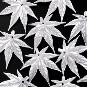 May include: A collection of eleven silver-colored maple leaf charms, each with a hole at the top for stringing. The leaves have detailed veining and are arranged on a black background.
