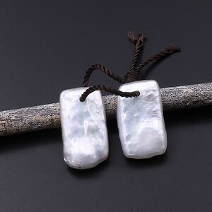 May include: Two white rectangular pearl pendants with brown cords. The pendants are irregular in shape and have a slightly iridescent sheen.