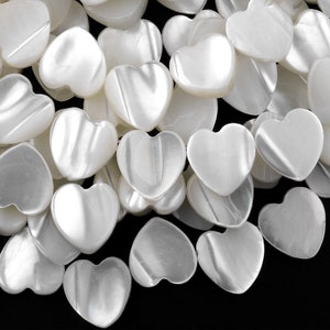 以下が含まれることがあります： 白いハート型のビーズの山。ビーズは真珠のような素材でできており、滑らかで光沢のある仕上がりです。