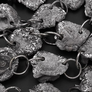 Könnte beinhalten: Eine silberne Kettenkette mit großen, unregelmäßig geformten, dunkelgrauen Metallgliedern. Die Glieder haben eine raue, strukturierte Oberfläche.