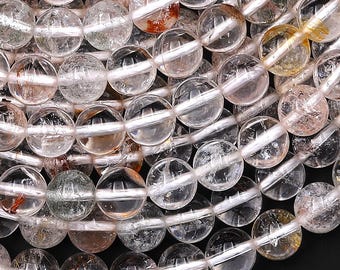 Puede incluir: Primer plano de un collar de cuentas de piedras preciosas de cuarzo transparente. Las cuentas son redondas y transparentes, con sutiles inclusiones y variaciones de color, incluyendo toques de rosa, amarillo y marrón. Las cuentas están ensartadas, creando un patrón orgánico y texturizado.