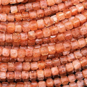 Può includere: Una collana di perle di pietra di sole arancione, ogni perla è di forma quadrata con una superficie leggermente sfaccettata. Le perle sono infilate insieme per creare una lunga collana o un braccialetto.