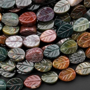 Puede incluir: Primer plano de un montón de cuentas de ágata ovaladas de color verde, marrón y rojo con un diseño de hoja tallado en cada cuenta.