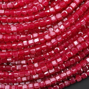 Może przedstawiać: Zbliżenie na kolekcję fasetowanych, kwadratowych koralików w głębokim czerwonym odcieniu. Koraliki są nawleczone, tworząc zakrzywiony wzór. Układ sugeruje naszyjnik lub podobną biżuterię, z naciskiem na kolor i fakturę.