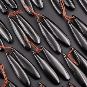 May include: A collection of black, polished, teardrop-shaped pendants with brown string ties. The pendants are arranged in rows on a dark surface.