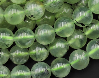 May include: A close-up of a strand of green aventurine beads with black inclusions. The beads are round and smooth, and they have a translucent appearance.