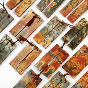 Puede incluir: Una colección de 18 colgantes rectangulares de piedra natural pulida con cordones de cuero marrón. Las piedras son una mezcla de colores marrón, verde y naranja con un patrón de mármol.