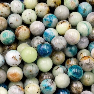 May include: A close-up of a pile of round, multi-colored gemstone beads. The beads are a mix of light blue, green, white, and brown. The beads are smooth and polished.
