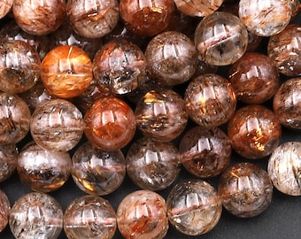 Può includere: Primo piano di una collana di perle rotonde, trasparenti, di colore marrone rossiccio con macchie dorate. Le perle sono in pietra naturale e hanno una finitura liscia e lucida.