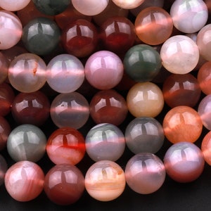 May include: A close-up of a collection of round, smooth agate beads in various shades of pink, red, brown, and green. The beads are polished and have a translucent appearance.
