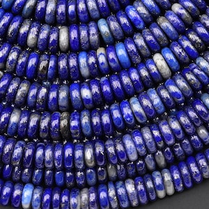 Puede incluir: Primer plano de una colección de cuentas de piedra preciosa de lapislázuli azul intenso. Las cuentas tienen forma de disco y tienen un acabado pulido y brillante. Algunas cuentas tienen inclusiones blancas y grises, lo que añade interés visual. Las cuentas están dispuestas en un patrón circular.