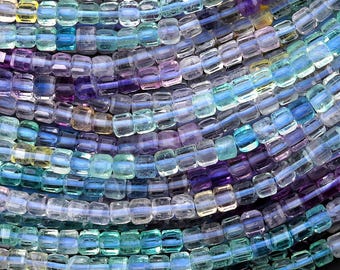 May include: Close-up of a collection of colourful beads in shades of blue, purple, green, and yellow. The beads are small, square-shaped, and strung together, creating a textured, layered effect. The beads are likely used for jewellery making or crafting.