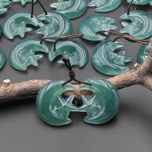 May include: Jade pendants in a crescent moon and star design, strung on brown cords. The pendants are a translucent teal green color. The pendants are arranged on a dark gray surface with a brown branch.