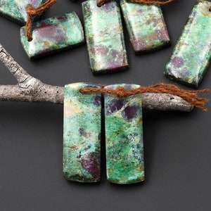 Op de afbeelding: Twee rechthoekige edelsteen hangers in groen, paars en bruin met een bruin touwtje.