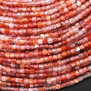 May include: Close-up of a collection of small, square-shaped beads in shades of red, orange, and pink. The beads are strung together, creating a layered, circular pattern. The beads have a faceted appearance, reflecting light.