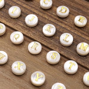 May include: A collection of white, round alphabet beads with gold-colored letters. The beads are arranged on a wooden surface, showcasing the letters A through Z. Ideal for jewelry making or craft projects.