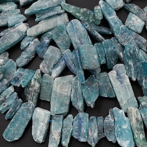 May include: A collection of blue-green kyanite gemstone beads, each with a unique, rough-hewn shape and a natural, striated texture. The beads are arranged in a random pattern, showcasing the variation in size and form.