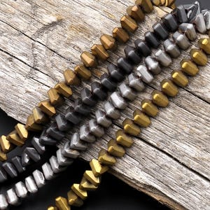 Puede incluir: Primer plano de varios hilos de cuentas metálicas en varios colores, incluyendo dorado, plateado y gris plomo. Las cuentas tienen una forma geométrica angular y están dispuestas sobre una superficie de madera envejecida.