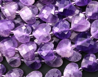 May include: Close-up of a collection of amethyst heart-shaped beads. The beads are a deep purple color with faceted surfaces, strung together with small, clear beads. The arrangement creates a textured, romantic aesthetic.