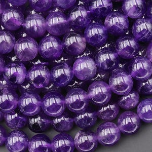 Puede incluir: Una colección de cuentas de amatista esféricas y lisas en varios tonos de violeta y morado intenso. Cada cuenta presenta un acabado pulido y brillante con inclusiones internas naturales y reflejos de luz en las superficies redondeadas.