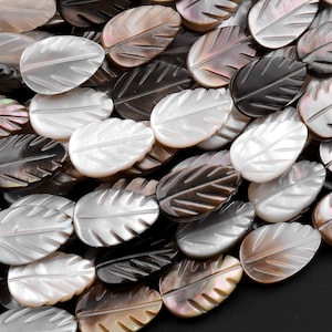 Puede incluir: Primer plano de un montón de cuentas ovaladas con forma de hoja. Las cuentas están hechas de nácar y tienen un patrón de color natural marrón, blanco y negro.