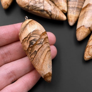 Könnte beinhalten: Eine Sammlung von braunen und beigefarbenen, gemusterten Steinanhängern mit einer silbernen Öse oben. Die Anhänger sind tropfenförmig und haben ein natürliches, organisches Aussehen.