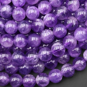 Puede incluir: Una colección de cuentas de amatista redondas y pulidas en varios tonos de púrpura y violeta intenso. Cada cuenta presenta inclusiones naturales y una superficie lisa y brillante, dispuesta en un grupo denso y superpuesto con reflejos visibles.