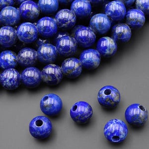 May include: A collection of round, polished lapis lazuli beads in varying shades of deep blue, with flecks of lighter blue and gold. Some beads have drilled holes, suggesting they are for jewelry making. The beads are arranged on a dark gray surface.