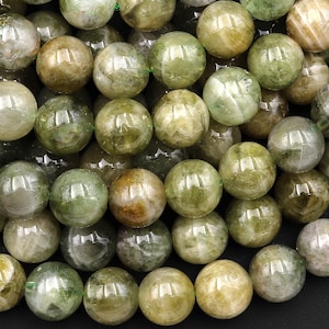 May include: A close-up of a collection of round, polished gemstones. The beads are primarily shades of green, with variations including olive, lime, and a hint of yellow. The beads are strung together on a green string.