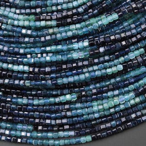 May include: Close-up of a collection of small, square-cut beads in shades of blue and teal. The beads are strung together, creating a layered, textured appearance. The colour gradient ranges from deep navy to light turquoise.