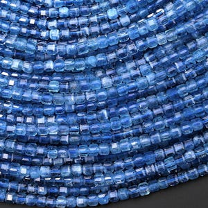 May include: Close-up of a collection of blue gemstone beads. The beads are small, square-cut, and strung together, creating a textured, layered effect. The color ranges from light to deep blue, with a slight sparkle.