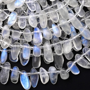 May include: Close-up of strands of oval moonstone beads. The translucent, milky white stones have flashes of blue and iridescent sheen. The beads are strung together, creating a shimmering, natural gemstone jewelry element.